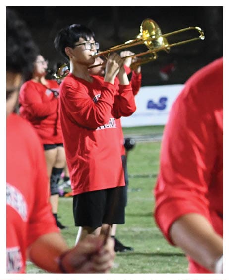 People wearing red shirts and black shorts are playing brass instruments on a grassy field, likely participating in a marching band practice or performance. Description generated by AI