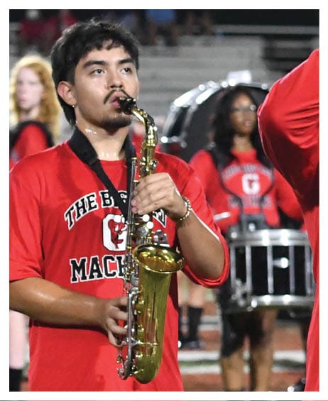 People wearing red shirts are playing musical instruments, including a saxophone and a drum, likely performing in a marching band on a field. Description generated by AI