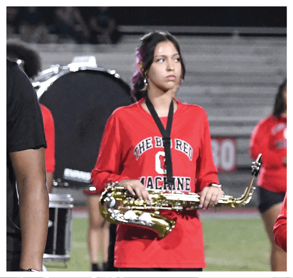 Two adults stand on a football field at night with one wearing a red polo and cap and the other in a black t-shirt while band members in red uniforms play instruments in the background. Description generated by AI
