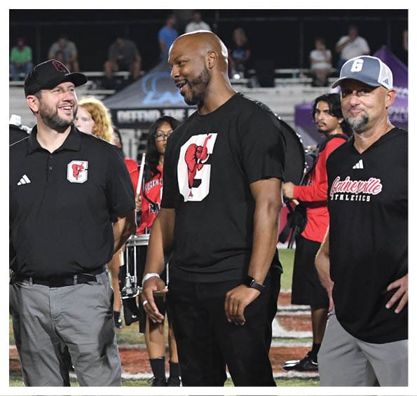 Four adults stand on a football field at night, smiling and holding hands, with some wearing red and black shirts featuring logos, and a band playing in the background. Description generated by AI