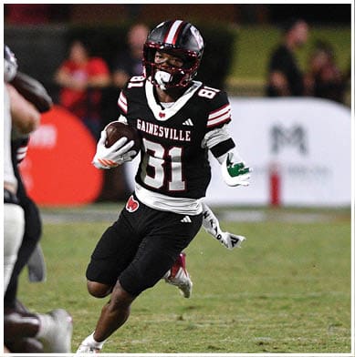 A person wearing a black and red Gainesville football uniform is running with the ball while being approached by a person in a white and blue uniform on a football field during a game. Description generated by AI
