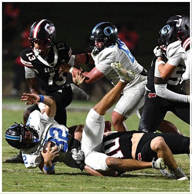 People in football uniforms are engaged in a play on a grassy field with one player in white and blue falling backward while others in black and red attempt to tackle or block. Description generated by AI