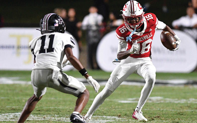 A person in a red and white football uniform is running with the ball while two people in white and black uniforms prepare to defend on a grassy field at night. Description generated by AI