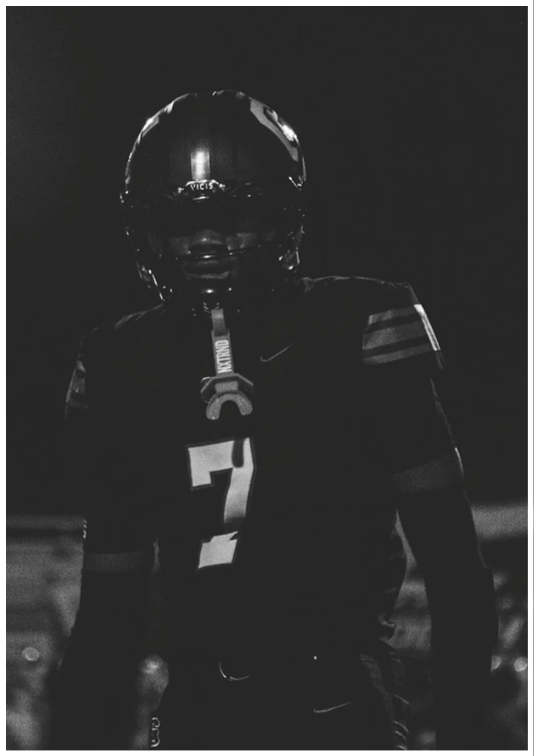 A person wearing a football uniform with the number seven stands in a dark setting, illuminated slightly from the front. Description generated by AI