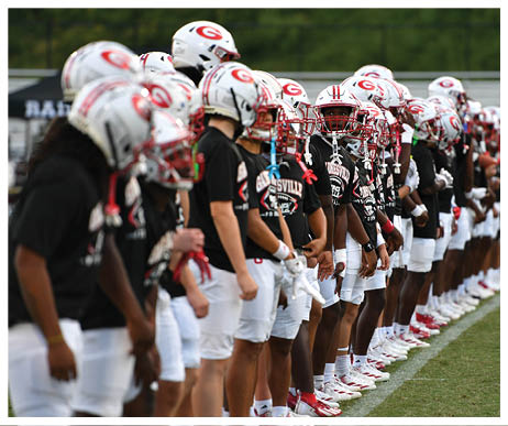 A line of people wearing football helmets, black shirts, and white shorts stand side by side on a grassy field. Description generated by AI