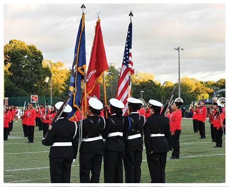 A group of people in uniform holding flags stands on a grassy field while a band dressed in red performs in the background during an outdoor event. Description generated by AI
