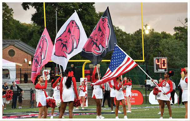 People in cheerleading uniforms hold flags including an American flag and team logos on a football field near a goalpost with a scoreboard in the background. Description generated by AI