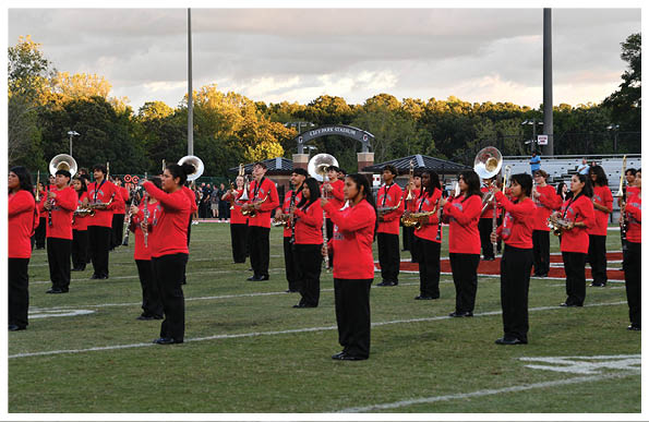 People wearing red jackets and black pants are standing on a football field playing brass and woodwind instruments in a marching band formation. Description generated by AI