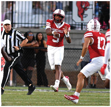 A group of people in football uniforms are playing on a field with one person in a red and white uniform throwing a football while others in red and white and black and white uniforms prepare to catch or defend. Description generated by AI