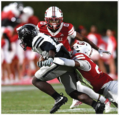 A person in a black and white football uniform is being tackled by a person in a red and white uniform on a football field with another person in a red and white uniform approaching in the background. Description generated by AI