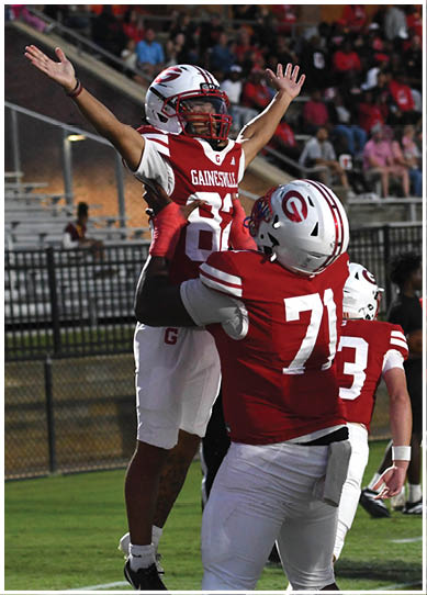Two people wearing red and white sports uniforms are embracing, with one person’s arm wrapped around the other’s back. Description generated by AI