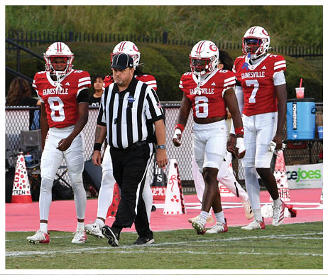 Three people in red and white football uniforms and a referee in black and white stripes walk across a football field with two other people standing near the sideline. Description generated by AI