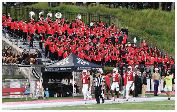A marching band dressed in red uniforms is seated in bleachers behind a football field where players in white and red uniforms and referees are walking on the sideline near a tent. Description generated by AI