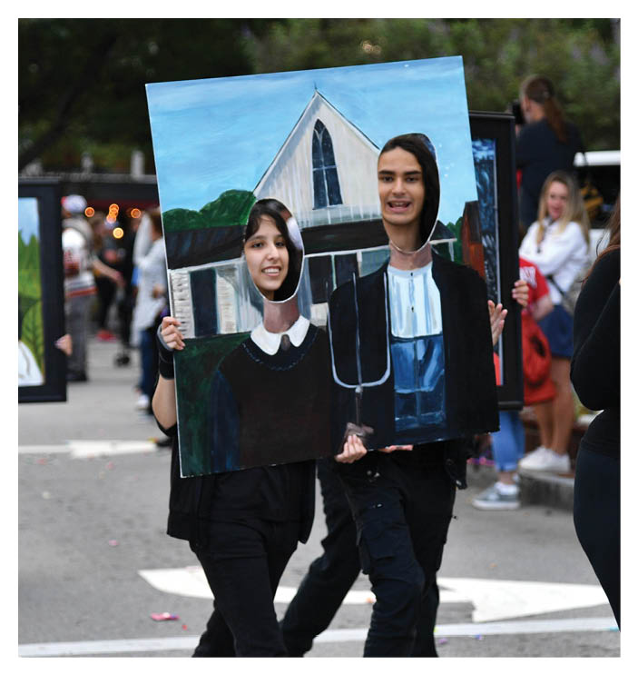 People hold painted boards with cutouts for faces depicting a parody of the American Gothic painting while walking on a street. Description generated by AI