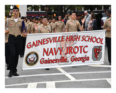 People in uniform march in a parade holding a banner that reads Gainesville High School Navy JROTC Gainesville, Georgia. Description generated by AI