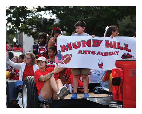 People wearing red and black cheerleading uniforms and casual clothing are on a float holding a banner that reads “Mund Mill Arts Academy" during an outdoor event. Description generated by AI
