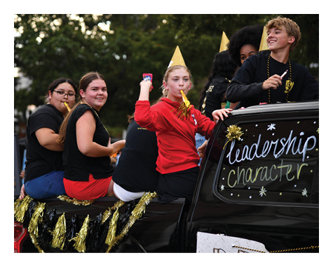People wearing party hats and festive clothing are sitting and standing on a decorated vehicle with words like leadership and character written on the window. Description generated by AI