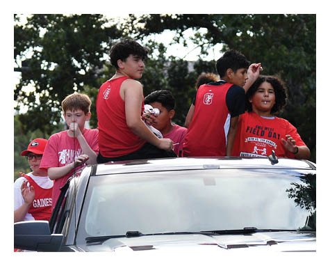 A group of people wearing red shirts are riding on and walking beside a black Chevrolet truck during an outdoor event with trees and spectators in the background. Description generated by AI