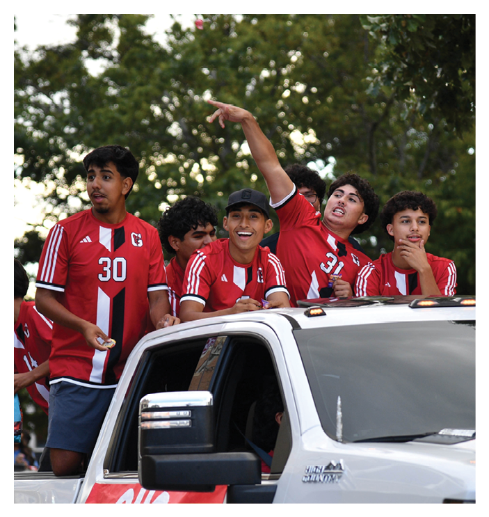 People wearing red sports jerseys are riding in the back of a white pickup truck with a red banner on the door during an outdoor event. Description generated by AI