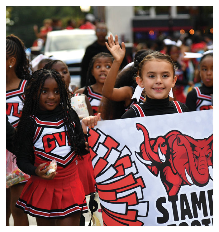 Children in cheerleading uniforms are holding a banner with a red elephant mascot and smiling during an outdoor event. Description generated by AI