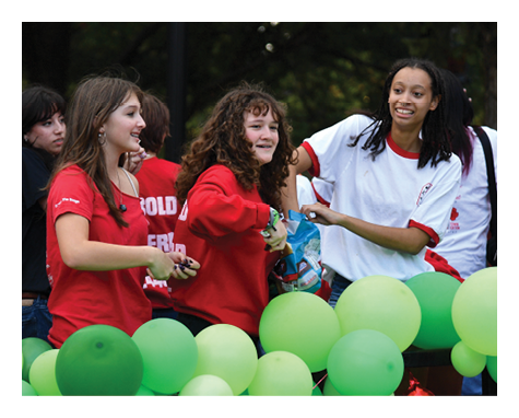 Three people wearing casual red and white shirts are smiling and interacting behind a decorative arrangement of green balloons. Description generated by AI