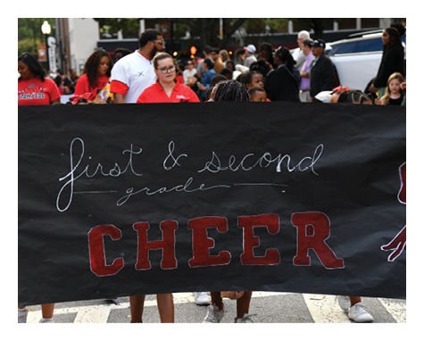 People wearing red shirts hold a large black banner that reads “first & second grade CHEER" during an outdoor event with a crowd in the background. Description generated by AI