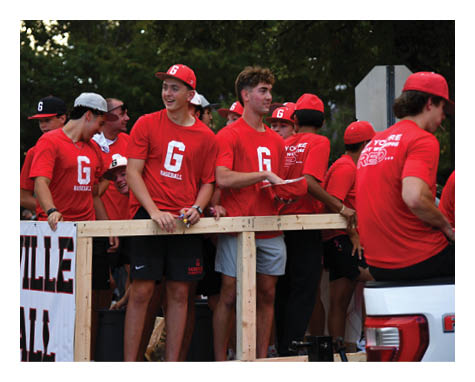 A group of people wearing matching red baseball shirts and caps are standing and sitting on a wooden platform attached to a vehicle outdoors. Description generated by AI