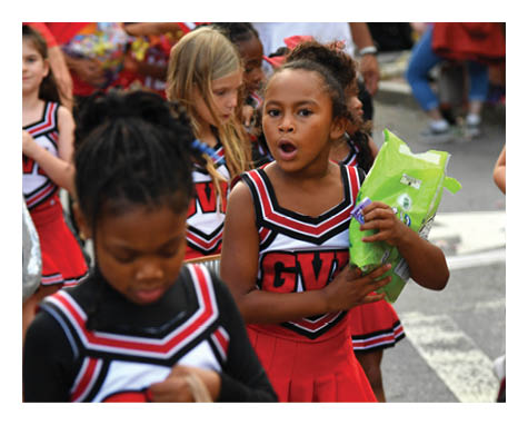 Children in red and black cheerleading uniforms are gathered outdoors, with one child holding a green bag and others holding various items. Description generated by AI