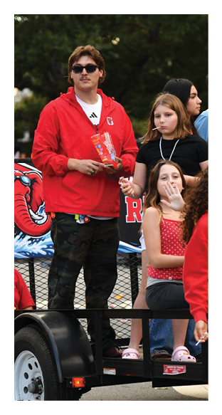 A group of children and an adult are gathered outdoors, some sitting and some standing, with a few holding snacks and wearing casual clothing and lanyards. Description generated by AI