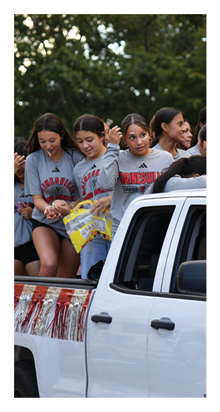 A group of people wearing matching long-sleeve shirts are sitting and standing in the back of a white pickup truck decorated with red and white streamers, appearing to enjoy a parade or outdoor event. Description generated by AI