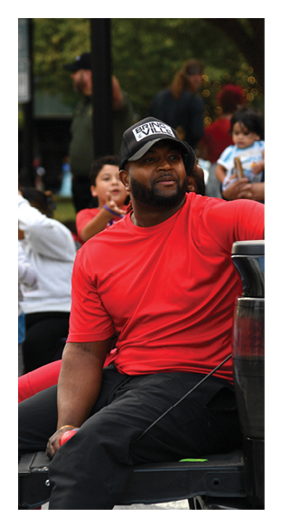 A person wearing a red shirt and a black and white cap is sitting on a bench while children and other people stand in the background at an outdoor event. Description generated by AI