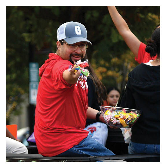 People wearing red shirts are sitting in the back of a black pickup truck, with one person smiling and handing out candy. Description generated by AI