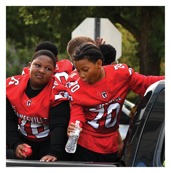 Children wearing red sports jerseys are gathered around a vehicle, with one child holding a water bottle and leaning on the vehicle. Description generated by AI