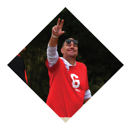 Two adults wearing red sports jerseys are outdoors, with one raising a hand and the other holding a wooden plank. Description generated by AI