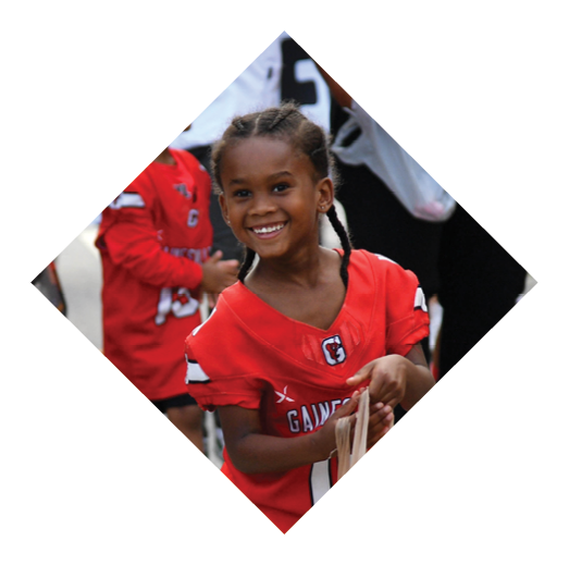 A child with braided hair wearing a red sports jersey is smiling and holding a bag while another child in a similar jersey stands in the background. Description generated by AI