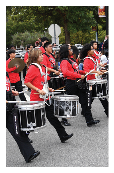 A group of people wearing matching red shirts and black pants are marching in formation while playing snare drums on a street. Description generated by AI