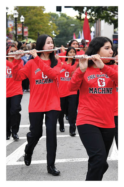 A group of people wearing matching red shirts and black pants are playing flutes while marching down a street, led by a person in a red shirt and beige pants. Description generated by AI