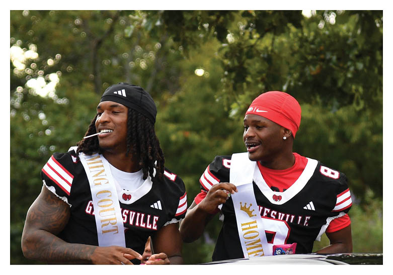 Two adults wearing black and red sports jerseys and sashes labeled “KING COURT" and "HOME" are smiling and interacting outdoors with trees in the background. Description generated by AI