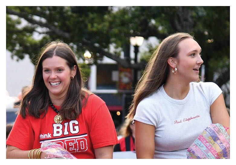 A person wearing a red shirt with the words “think BIG RED" is smiling and holding a colorful bag while sitting on a vehicle in a parade with other vehicles and people in the background. Description generated by AI