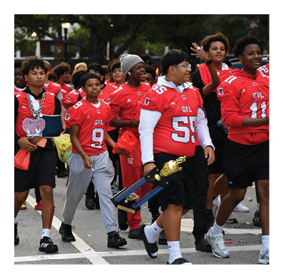 A group of people wearing red sports jerseys are walking together outdoors, with one person holding a large trophy and others smiling and interacting. Description generated by AI