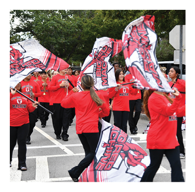People wearing red shirts are marching outdoors while playing instruments and waving large red and white flags with text and graphics. Description generated by AI