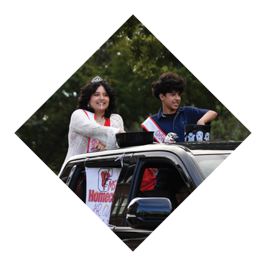 Two people wearing sashes and smiling are standing in a vehicle decorated with a homecoming sign during an outdoor event. Description generated by AI