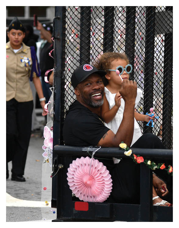 A person wearing a hat and black clothing is sitting on a decorated float with flowers and paper decorations during an outdoor event near a banner that partially reads “HIGH SCHOOL" and "Georgia." Description generated by AI