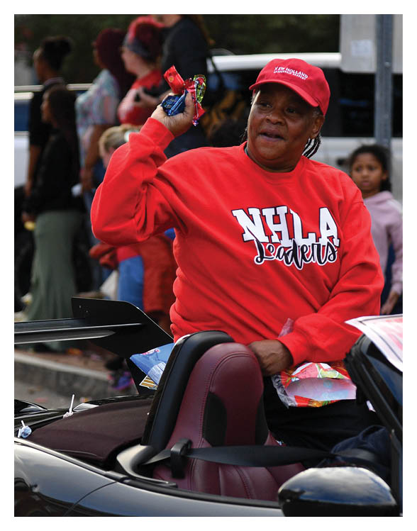 A person wearing a red NHLA Leaders sweatshirt and red cap is sitting in a convertible car and throwing candy to a crowd of people standing nearby. Description generated by AI