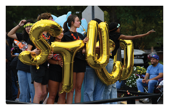 A group of people standing outdoors hold large gold balloons spelling out “STU" while others sit nearby on chairs. Description generated by AI