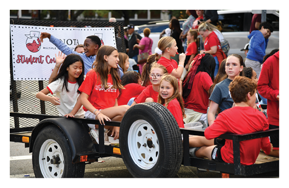 A group of children wearing red and casual clothing are sitting and standing on a trailer in an outdoor setting, appearing to be part of a student council event. Description generated by AI