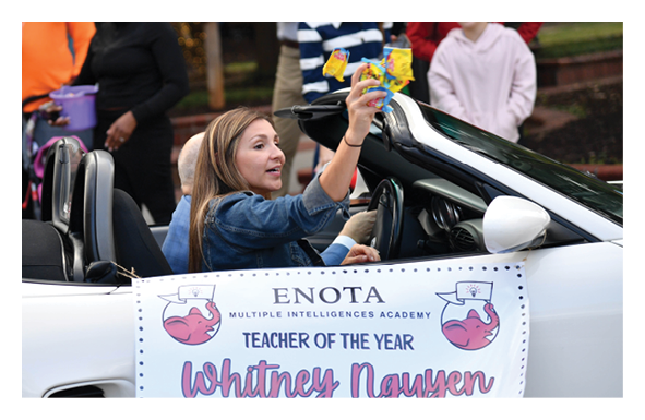 A person with long hair wearing a denim jacket is sitting in a white convertible car holding up candy while a banner on the car reads “ENOTA Multiple Intelligences Academy Teacher of the Year Whitney Nguyen." Description generated by AI