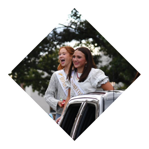 Two adults wearing sashes stand and smile on the back of a vehicle in a parade with other people and vehicles visible in the background. Description generated by AI