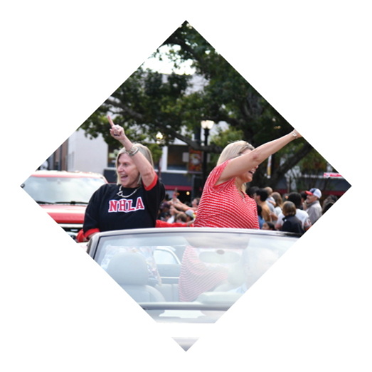 Two adults smiling and waving while standing in a convertible car during a parade with a crowd in the background. Description generated by AI