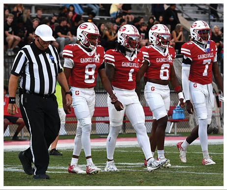 Four football players in red and white uniforms walk side by side on a field between two referees wearing black and white striped shirts with a crowd in the background. Description generated by AI